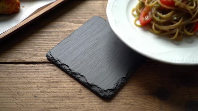 Slow motion footage of a black slate coaster next to a plate of spaghetti pasta on a rustic wooden table.