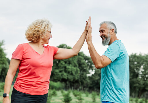 Smiling active mature mid aged couple jogging exercising and having fun and giving high five together taking a break in the park - Powered by Adobe