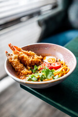 A bowl of spicy tempura shrimp ramen with noodles, soft-boiled egg halves, cherry tomatoes, and fresh cilantro in a rich, vibrant broth, served on a green table
