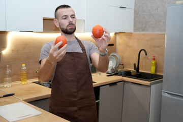 In a cozy kitchen, a male chef in an apron carefully examines two fresh tomato vegetables, considering how to use them in dinner preparation. The atmosphere is both focused and inviting