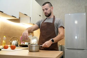A man in an apron cooks in a stylish, well-equipped kitchen. He pours grains into a saucepan, surrounded by fresh vegetables and kitchen tools
