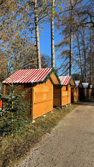 A line of warm wooden Christmas Market huts with red-and-white striped roofs, decorated with garlands, set along a sunlit forest path. Ideal for festive markets, winter scenes, and holiday visuals.