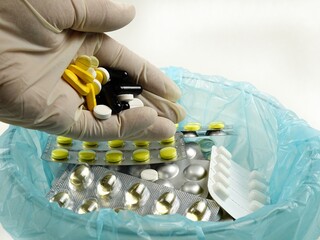 A hand in a white rubber glove pours expired pills into a container with expired pill blister packs medicines

