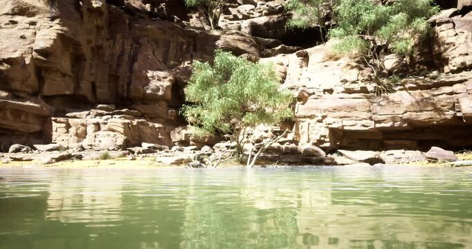Lush greenery thrives next to tranquil water, reflecting the rocky cliff formations in the background. The setting is illuminated under clear blue skies, creating a peaceful atmosphere.