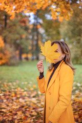 A young woman holds a maple leaf in her hand in a colorful autumn park. Golden, cozy autumn concept.
