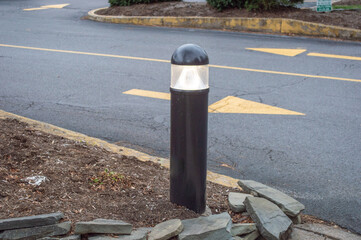 Modern black bollard landscape light illuminated near driveway with yellow road arrows and stone retaining wall.