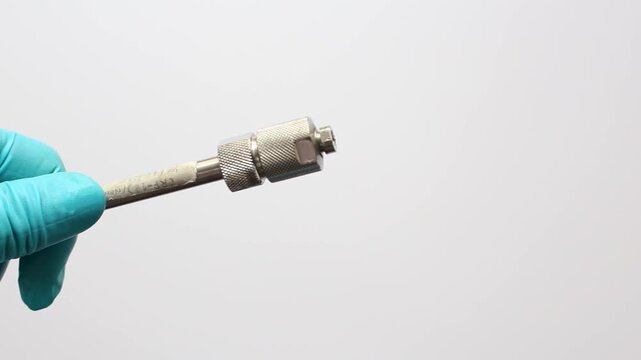 A scientist's hands in blue gloves connect a stainless steel capillary fitting to an HPLC column for liquid chromatography analysis 
in a science lab.
