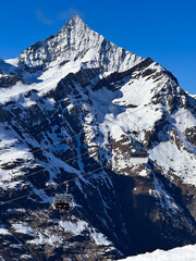 Snowy mountain peak with cable cars ascending the steep slopes in the Swiss Alps. Clear blue sky, dramatic winter landscape and alpine scenery captured in crisp detail.