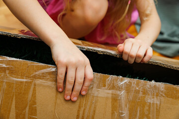 Close-up of a child’s hands opening a cardboard box, lifting the lid after peeling tape, captured in a natural home setting.