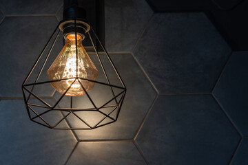 A modern geometric pendant lamp hangs against a dark tiled wall. Warm Edison bulb