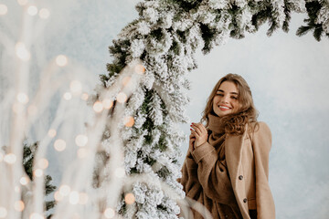 Happy young woman in a cozy camel coat and chunky knit turtleneck smiling brightly under a large snowy fir garland arch in a winter studio