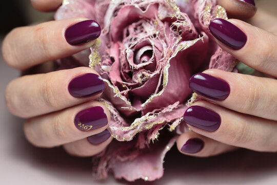 Nails are covered gel polish with the golden inscription "love". Soft hands with glossy purple nails gently cradle a textured rose. The nails exhibit a lovely shine, adding elegance to the scene.
