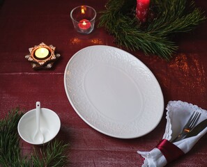 Christmas table setting on a burgundy tablecloth with a white oval platter, candles, and a napkin.