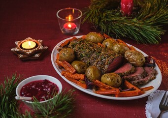 A cooked roast beef with carrots, onions, and potatoes on a white platter on a festive Christmas table setting. Merry Christmas! Beef dishes.
