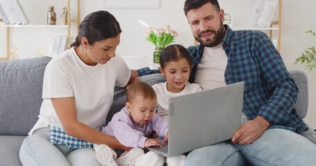 Happy family uses laptop online at home. Parents and children sit on the sofa, smiling and browsing the screen together while a baby taps keys. Concept: modern family time online at home. - Powered by Adobe
