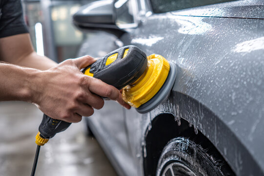 Man using electric polishing machine to clean and shine wet silver car surface during detailed vehicle maintenance indoors