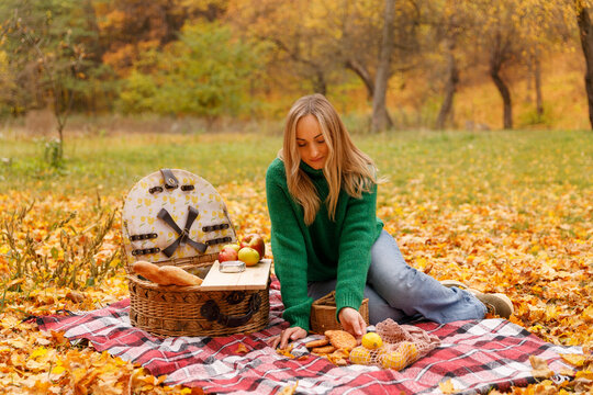 Mid-adult woman sitting on a red plaid blanket surrounded by yellow leaves, reaching into a wicker picnic basket containing fruit and bread. Enjoying a relaxing autumn meal. - Powered by Adobe