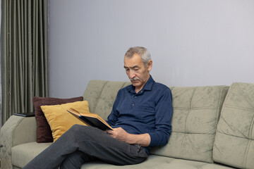 Fototapeta premium An elderly Asian man reading a book while sitting on the sofa at home