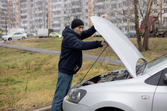 An elderly man inspects a car for serviceability by opening the hood. A broken car in winter.