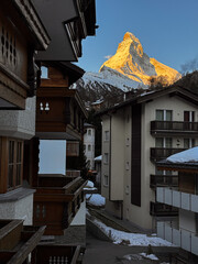 Scenic sunrise view of the Matterhorn rising between traditional Swiss alpine buildings in Zermatt, Switzerland. Morning light, winter season, peaceful mountain village atmosphere.