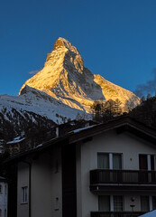 Scenic sunrise view of the Matterhorn rising between traditional Swiss alpine buildings in Zermatt, Switzerland. Morning light, winter season, peaceful mountain village atmosphere.