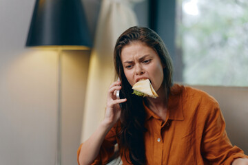 Young woman with long hair, wearing an orange shirt, multitasking as she talks on the phone while eating a sandwich in a cozy indoor setting