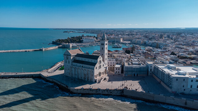 A panoramic coastal view of Trani shows the famous seaside cathedral overlooking turquoise waters, a long breakwater with lighthouses, and the historic old town bathed in bright Mediterranean sunlight