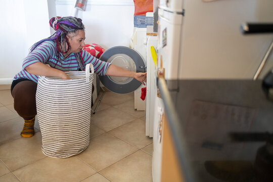 Plus size woman with dreadlocks doing laundry, reaching into a washing machine door next to a large laundry basket. - Powered by Adobe