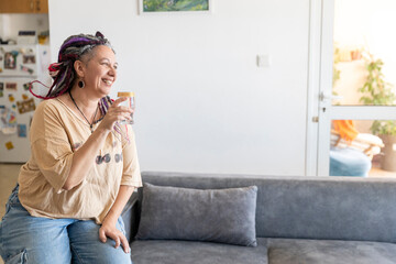 Happy plus size woman with dreadlocks sitting on a sofa in her living room, enjoying a refreshing drink.