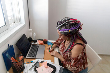Plus size woman with dreadlocks sitting at a cluttered desk, writing a note next to her laptop and...