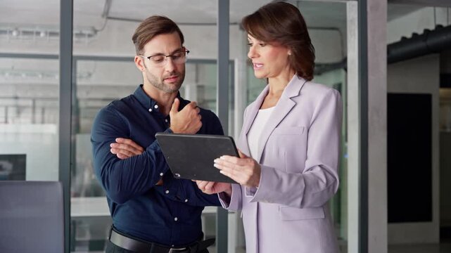 Mature Latin business woman and European young man discussing finance project on tablet gadget in office. Two diverse partners team, professional business people mentor and intern working together