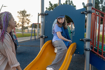 Fototapeta premium Plus size woman with dreadlocks standing next to a young girl sitting on an orange slide at a playground, looking at her.