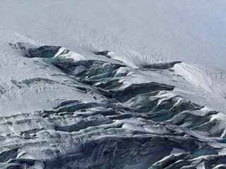Aerial view of dramatic snowy mountain textures and glacier crevasses in winter light. Natural abstract patterns created by ice, wind, and snow on a high-altitude alpine slope.