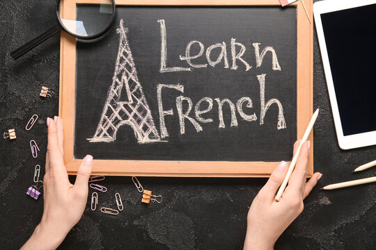 Female hands, chalkboard with text LEARN FRENCH, stationery and tablet computer on dark background, closeup - Powered by Adobe