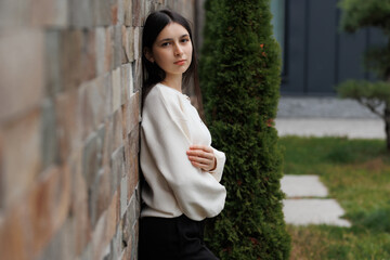 Young woman leans against a stone wall in a garden setting during the day, showcasing a relaxed and contemplative mood