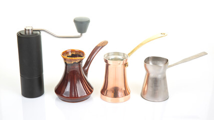 A black manual coffee grinder with a metallic handle stands beside a ceramic cezve, a copper cezve...