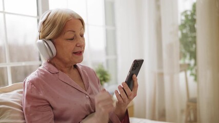Cheerful mature woman with headset listening to music on phone, resting in bed - Powered by Adobe