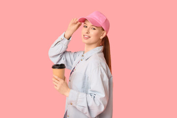 Beautiful young woman in stylish cap with paper cup of coffee on pink background