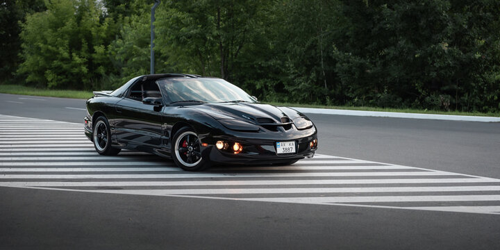 black Pontiac Firebird Trans Am parked on a painted traffic island. Wide three quarter front view of classic 1990s american pony car on empty rural road in a green forest. Ukraine - July 26 2025