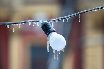 A single light bulb encased in ice, with icicles forming on a wire, set against a soft, colorful bokeh background, symbolizing the chill and beauty of a winter day.