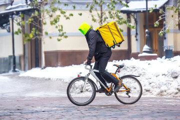 A food delivery courier in a neon green hoodie and yellow backpack rides a bicycle on a snowy urban street. Snow-covered buildings and trees are in the background, capturing a winter city scene.