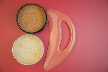 Two jars of natural body scrubs, one brown sugar and one cream, placed beside a pink massage tool on a vibrant red background, showcasing skincare products and wellness