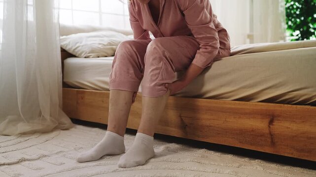 Elderly woman rubbing swollen legs while sitting on bed, varicose veins, cramps
