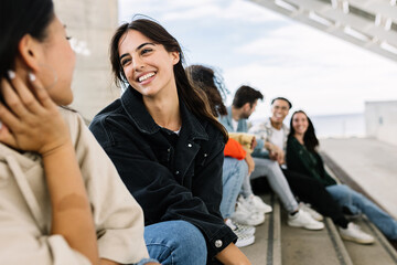 Portrait of two young multiracial women hanging out sitting outdoors with diverse friends. Friendship and bonding concept