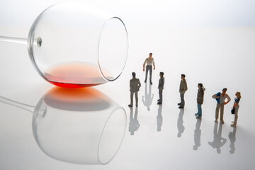 Six miniature figurines stand before a giant wine glass with red wine, symbolizing celebration or temptation. Reflective white background enhances the minimalist, conceptual design
