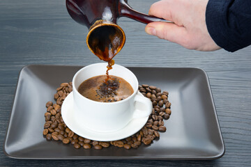 A close-up of a hand pouring rich Turkish coffee from a cezve into a cup, with coffee beans scattered around on a sleek black tray, framed by a warm wooden background.