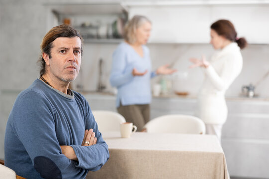 Thinking man sits in kitchen and in background adult daughter quarrels with elderly mother