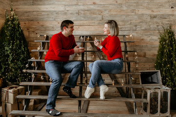 Happy young couple in matching red sweaters sitting on wooden steps, chatting and drinking hot cocoa in a rustic Christmas setting