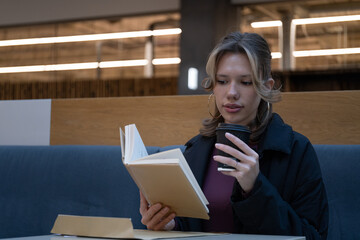 Fototapeta premium Young woman reads a book while holding a takeaway coffee cup in a cafe setting