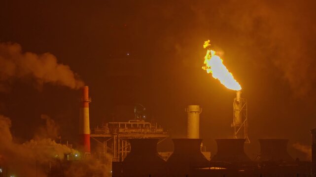 An industrial flare stack burns off excess gas with a large, bright orange flame and thick smoke against a dark night sky. Slow motion footage.